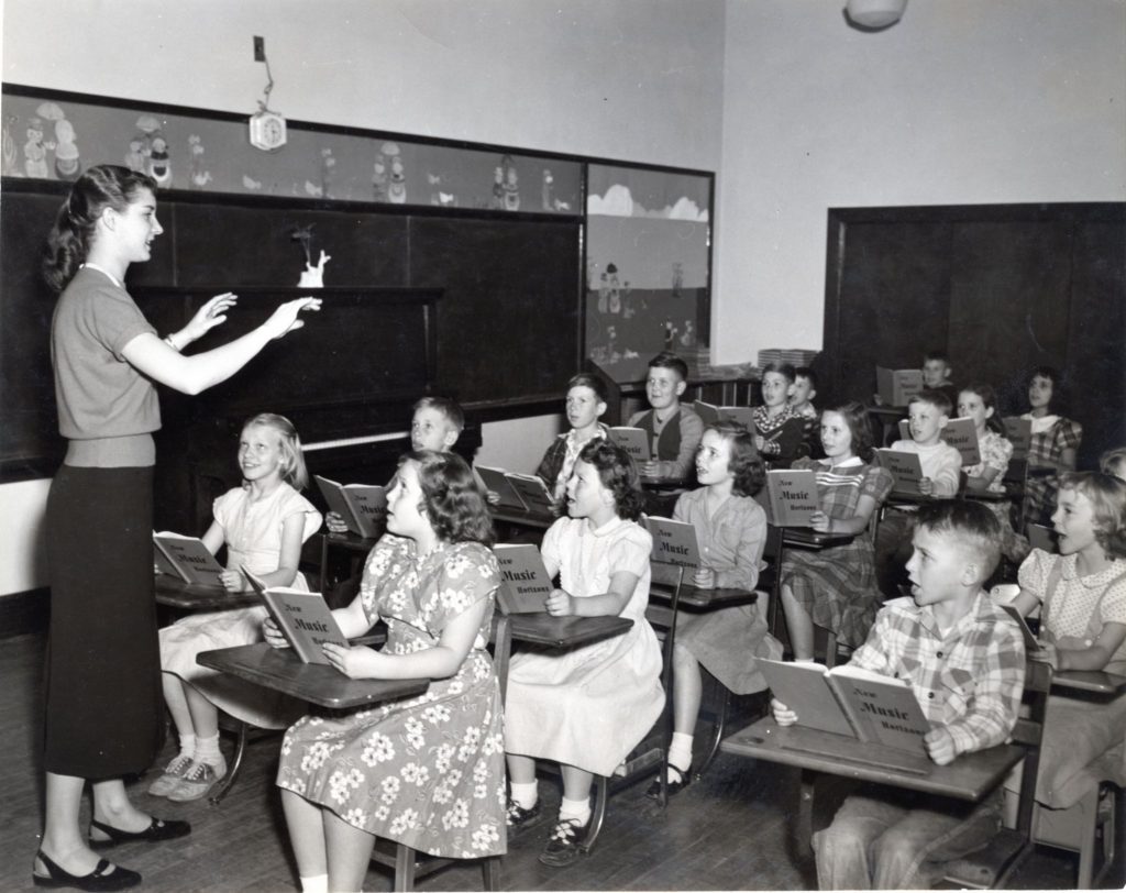 Black-and-white photo of a classroom where a female teacher stands at left, conducting with raised hands. About twenty elementary-age children sit in wooden desks holding songbooks titled New Music Horizons and appear to be singing. Behind them are chalkboards and a decorated bulletin board; a wall clock hangs high. A piano rests against the back wall. The students wear mid-20th century clothing such as dresses, sweaters, and plaid shirts. The room has hardwood floors and bright overhead lighting.