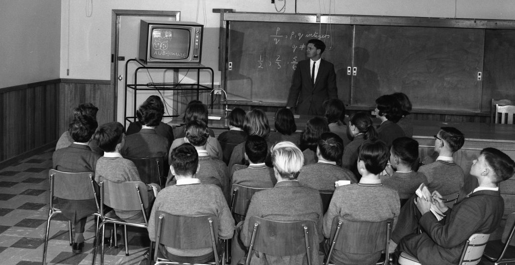 Black and white photo of a midcentury classroom. Around two dozen students sit on simple wooden chairs facing the front. Most wear sweaters and collared shirts, some with ties, and hold notebooks. At the front a male teacher in a suit stands beside a long chalkboard with math, including 1/2, 2/3, 5/6 and p, q notation. A small television on a wheeled cart shows a chalkboard graphic with set notation A U B. The room has checkered linoleum floor, wood wall paneling, and a lab bench with a sink.