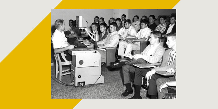 Black and white photograph of a classroom lecture. An instructor sits at the left beside a large, boxy audiovisual machine on wheels with cables, dials, and a small CRT monitor mounted on an arm. Rows of students, mostly young men, sit in tablet arm chairs facing forward, some taking notes. The scene suggests a mid 20th century setting with formal attire. The room has plain walls and a textured floor. The image is presented within a design frame featuring gray background and yellow diagonal shapes at two corners.