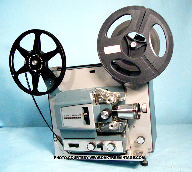 A product photo of a vintage Bell & Howell film projector against a light blue background. Two reels are mounted: a black take-up reel on the left marked Autoload and a larger gray reel on the right with a small white label. The projector has a blue gray metal housing, a prominent lens, film guides and rollers, and a front control panel with knobs and switches. A power cord drapes from the side. A caption at the bottom reads Photo courtesy www.oaktreevintage.com.