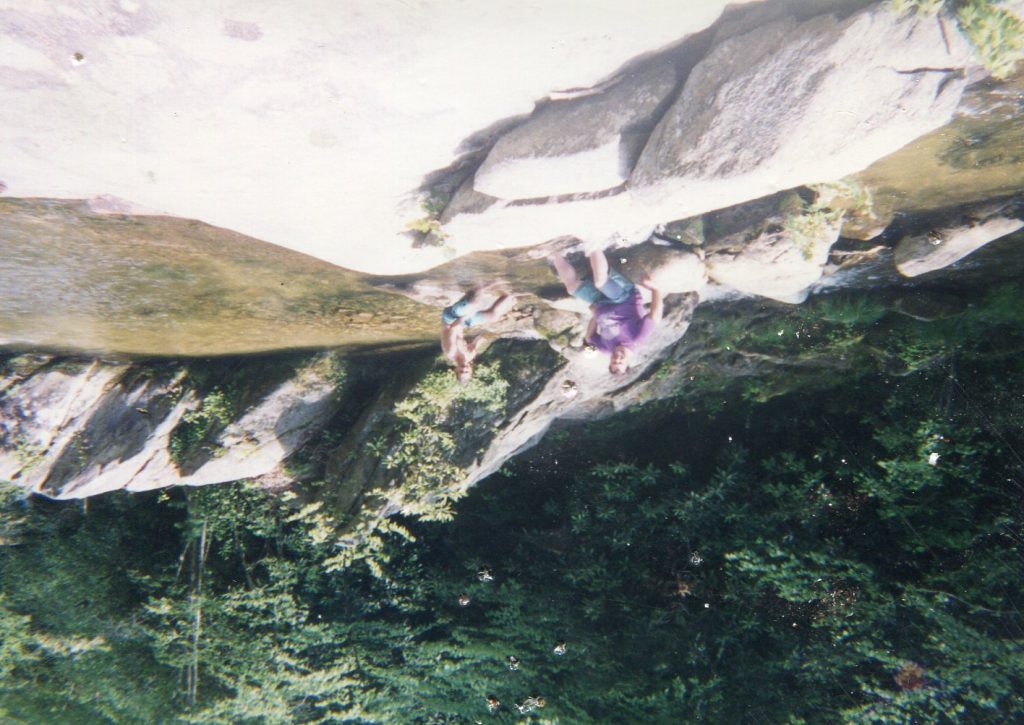 An outdoor scene at a shallow rocky stream in a forest. Two people sit on boulders by the water. The person on the left wears a purple shirt and shorts with feet in the stream. The person on the right is shirtless in shorts, sitting in the water. Large smooth rocks fill the foreground, and a sloped rock face and dense green trees line the background. The water is clear and calm. The photo appears slightly faded with small specks, like an old snapshot.