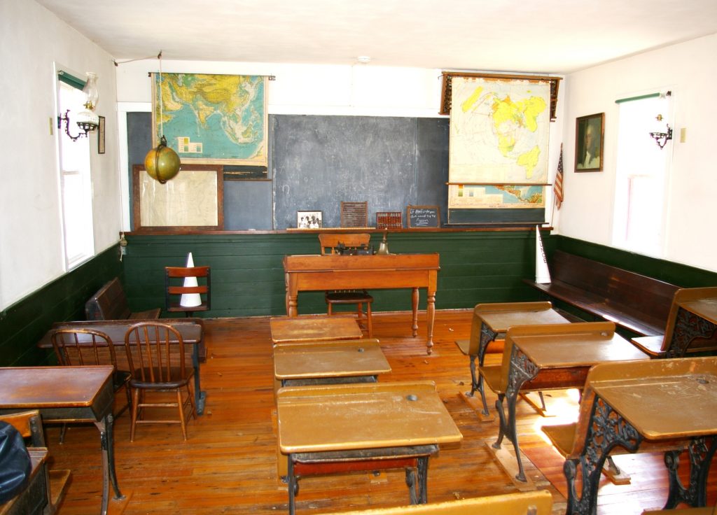 An old fashioned one room classroom with wooden floor and rows of cast iron and wood student desks facing a central teachers desk. A hand bell sits on the desk with a small stool beneath. A large chalkboard spans the wall, flanked by pull down world maps and a hanging split globe model. Shelves hold slates, an abacus, and a small photo. Long wooden benches line the sides. Sunlit windows, wall oil lamps, a framed portrait, and a U.S. flag complete the room.