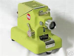 A retro, lime green tabletop optical device on a light surface. It has a cylindrical lens housing on the front right with a knurled focus ring, several small metal levers and knobs, and a ventilated top panel. A red circular badge with the letters as appears on the side. At the front base is a metal slot or carrier, likely for inserting a slide or film frame. The unit stands on small feet and looks like a compact slide or film viewer or projector.