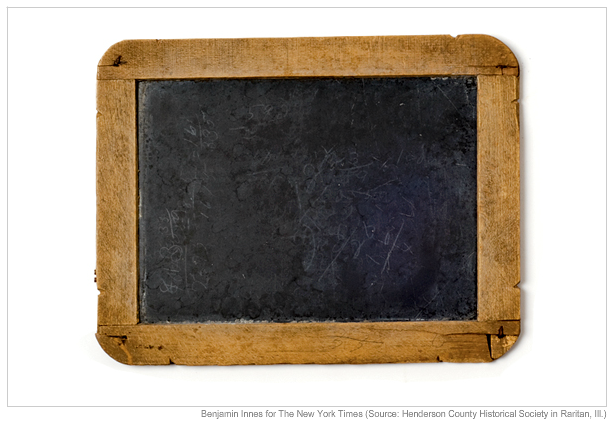 Photo of a small rectangular slate chalkboard with a thick light brown wooden frame on a white background. The slate surface is dark gray and worn, with faint erased chalk marks and scratches. The frame has rounded corners, visible joints, and small metal pins, showing age and slight chips. Lighting is bright and even, creating soft shadows around the board. No text, people, or additional objects are present. It is not a document scan.