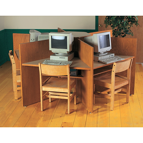 A classroom or library computer pod with a four way wooden study carrel on a light wood floor. Each quadrant holds an old beige CRT monitor, keyboard, and desktop tower, with a simple wooden chair pulled up to the desk. The monitors sit on risers and the dividers provide privacy around each station. In the background are green wall trim, a door with a brass knob, a whiteboard or screen, and a potted plant. The overall scene suggests a school setting from the late 1990s or early 2000s.